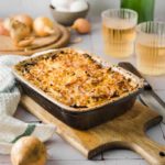 Auflaufform mit gebackenem Zwiebelgericht auf Holzbrett, daneben Zwiebeln und zwei gefüllte Gläser.