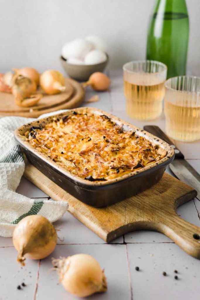 Auflaufform mit gebackenem Zwiebelgericht auf Holzbrett, daneben Zwiebeln und zwei gefüllte Gläser.