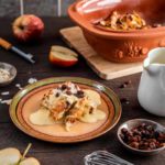 Apfel-Rosinen-Dessert mit Vanillesoße auf einem Teller, umgeben von Zutaten und Kochutensilien auf Holztisch.