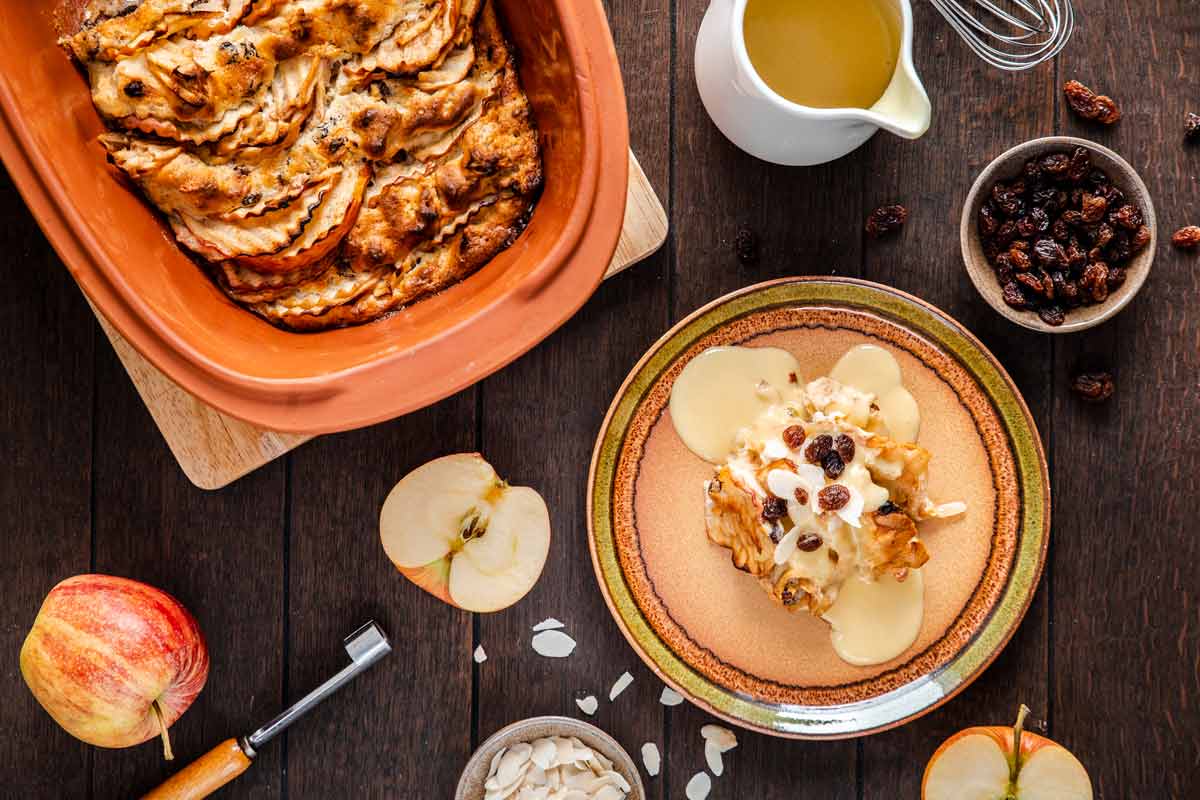 Apfel-Brotauflauf mit Vanillesauce, Rosinen und Mandeln auf Teller, umgeben von Äpfeln, Rosinen und einem Krug Sauce.