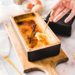 Hand löffelt Crème Brûlée aus rechteckiger Form auf Holzbrett, mit Vanilleschote als Deko.