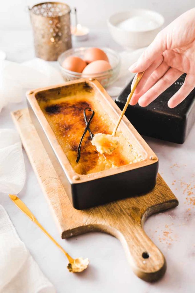 Hand löffelt Crème Brûlée aus rechteckiger Form auf Holzbrett, mit Vanilleschote als Deko.
