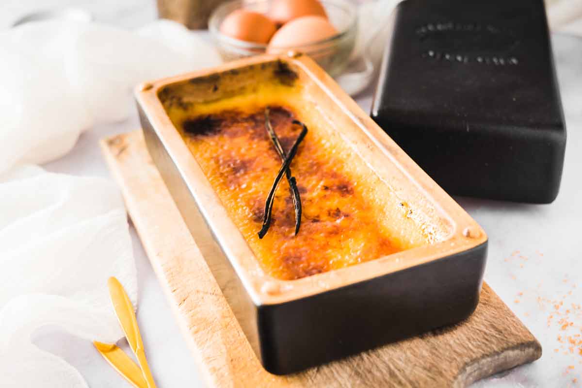 Crème Brûlée in einer rechteckigen Form mit Vanilleschote serviert, auf einem Holzbrett, im Hintergrund Eier.