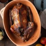 Gebratene Gans im Bräter, umgeben von Rotkohl, Soße, Kartoffeln und Äpfeln auf einem Holztisch.