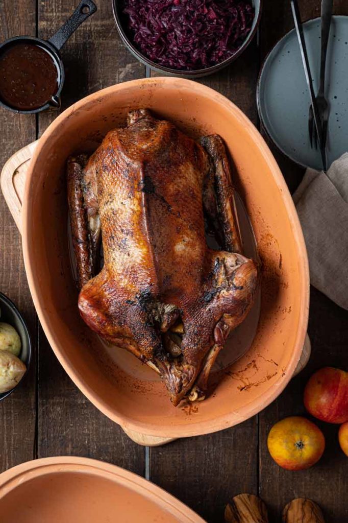 Gebratene Gans im Bräter, umgeben von Rotkohl, Soße, Kartoffeln und Äpfeln auf einem Holztisch.