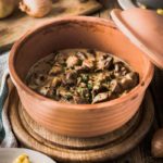 Ton-Schüssel mit Pilzragout in Sahnesoße, daneben Teller mit Spätzle, Holzbrett mit Zwiebeln im Hintergrund.