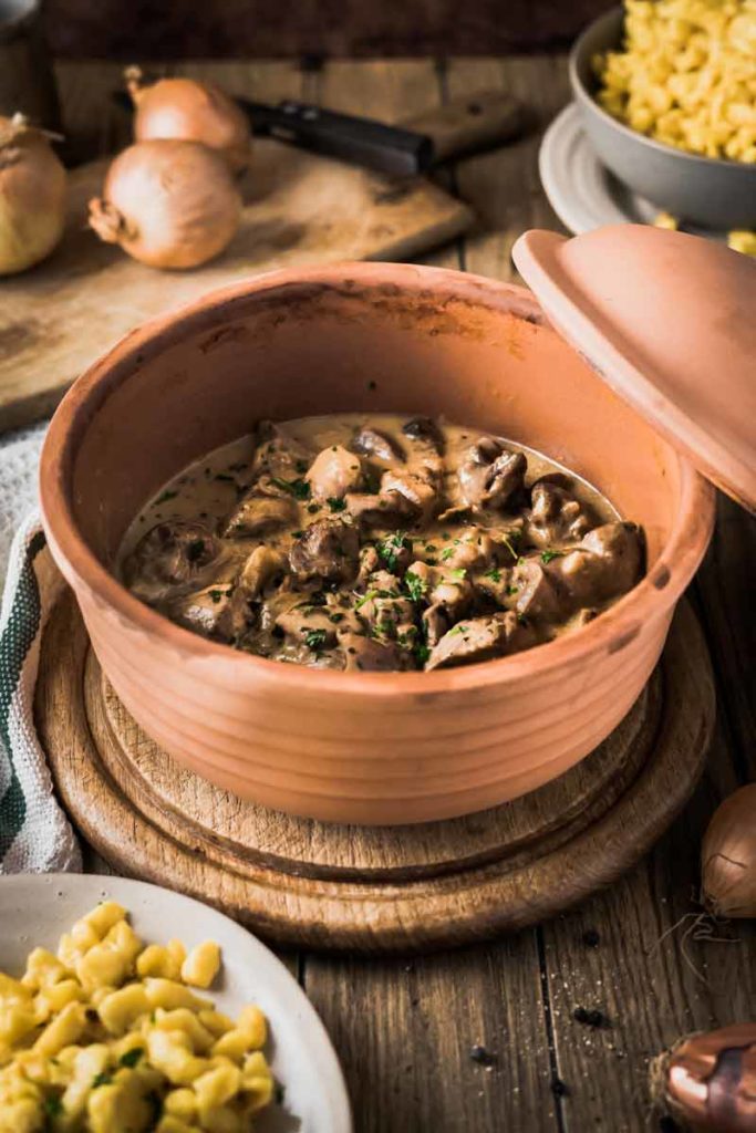 Ton-Schüssel mit Pilzragout in Sahnesoße, daneben Teller mit Spätzle, Holzbrett mit Zwiebeln im Hintergrund.
