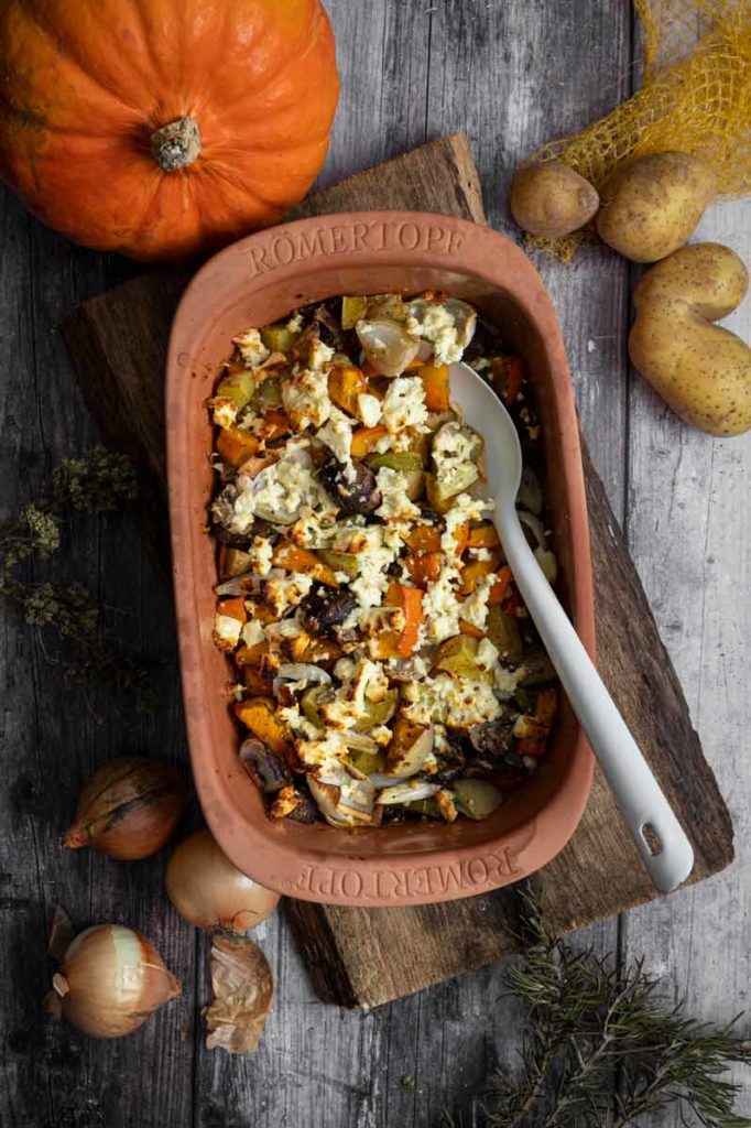 Auf einem Holztisch steht ein Römertopf mit Kürbisauflauf, umgeben von Kürbis, Kartoffeln und Zwiebeln.