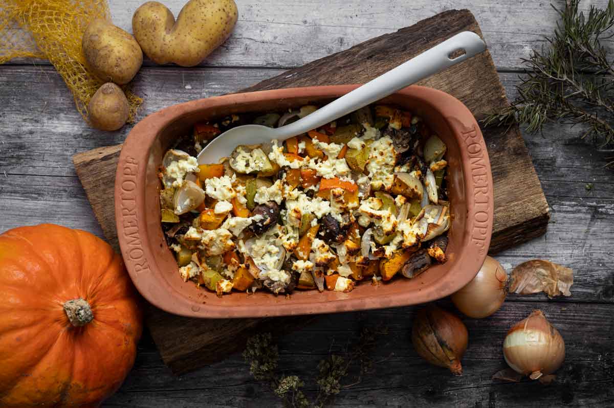 Auflaufform mit gebackenem Gemüse und Feta, daneben Kürbis, Kartoffeln und Zwiebeln auf rustikalem Holztisch.