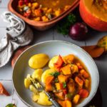 Teller mit Kartoffeln und Kürbiseintopf, garniert mit frischen Kräutern. Römertopf und halber Kürbis im Hintergrund.