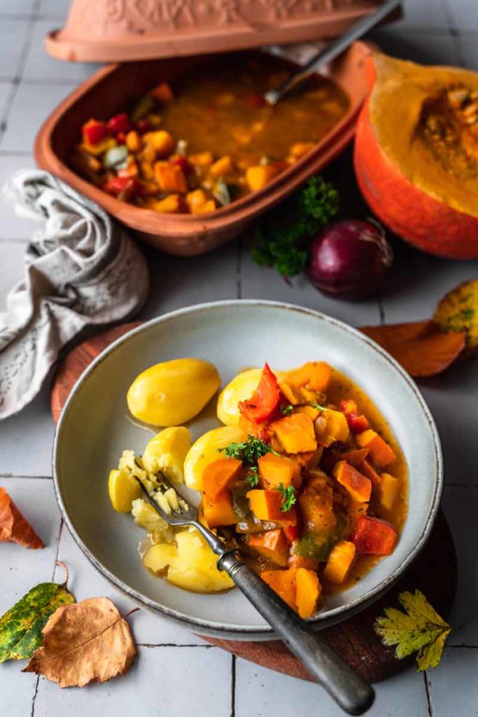Teller mit Kartoffeln und Kürbiseintopf, garniert mit frischen Kräutern. Römertopf und halber Kürbis im Hintergrund.