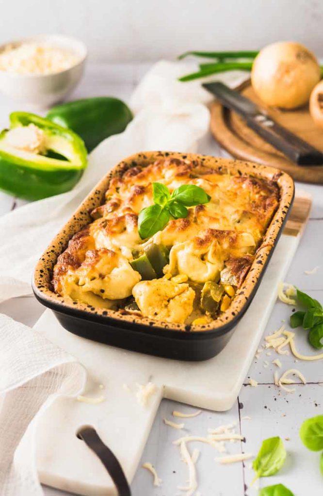 Überbackene Pasta mit Gemüse und Käse in einer Auflaufform, dekoriert mit Basilikum, neben Paprika und Zwiebel.
