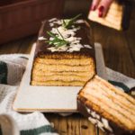 Rechteckige Torte mit Schichten, Schokoglasur, Mandelsplittern und Rosmarin dekoriert, auf Tisch mit Hand im Hintergrund.