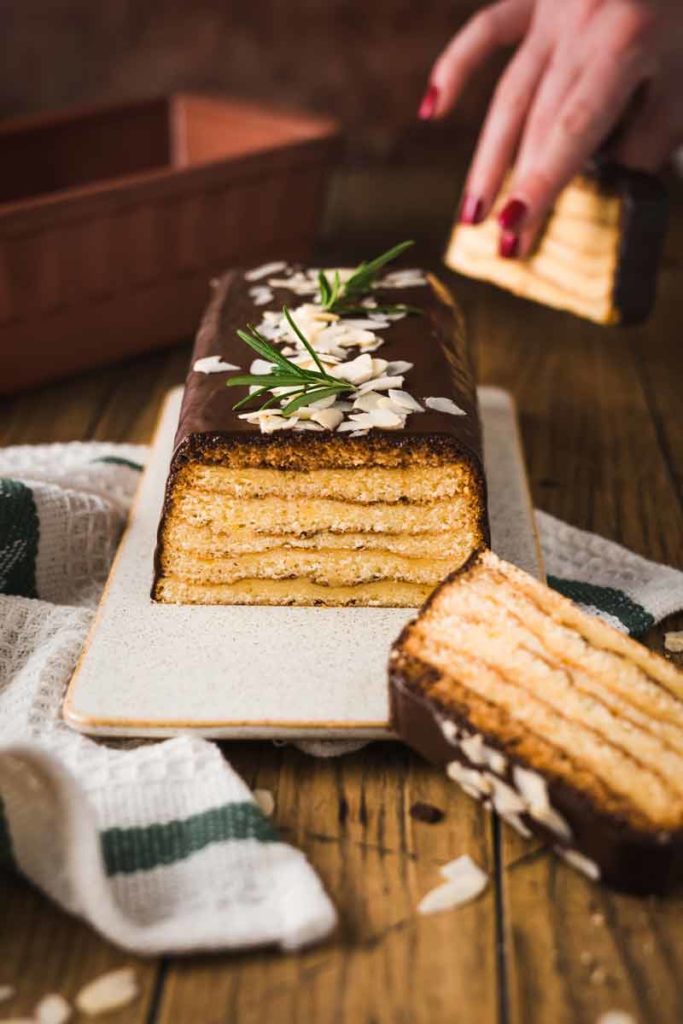 Rechteckige Torte mit Schichten, Schokoglasur, Mandelsplittern und Rosmarin dekoriert, auf Tisch mit Hand im Hintergrund.