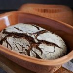 Roggenbrot mit knuspriger Kruste im runden Tontopf auf Holzbrettern, dunkelgrauer Hintergrund.