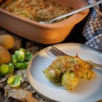 Auf einem Holztisch steht eine Auflaufform mit Rosenkohlgratin, daneben ein Teller mit einer Portion und ein Besteck.