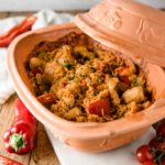 Keramiktopf mit Paprika-Hähnchen-Reis-Gericht, umgeben von frischen Paprikas und Tomaten auf Holzbrett.