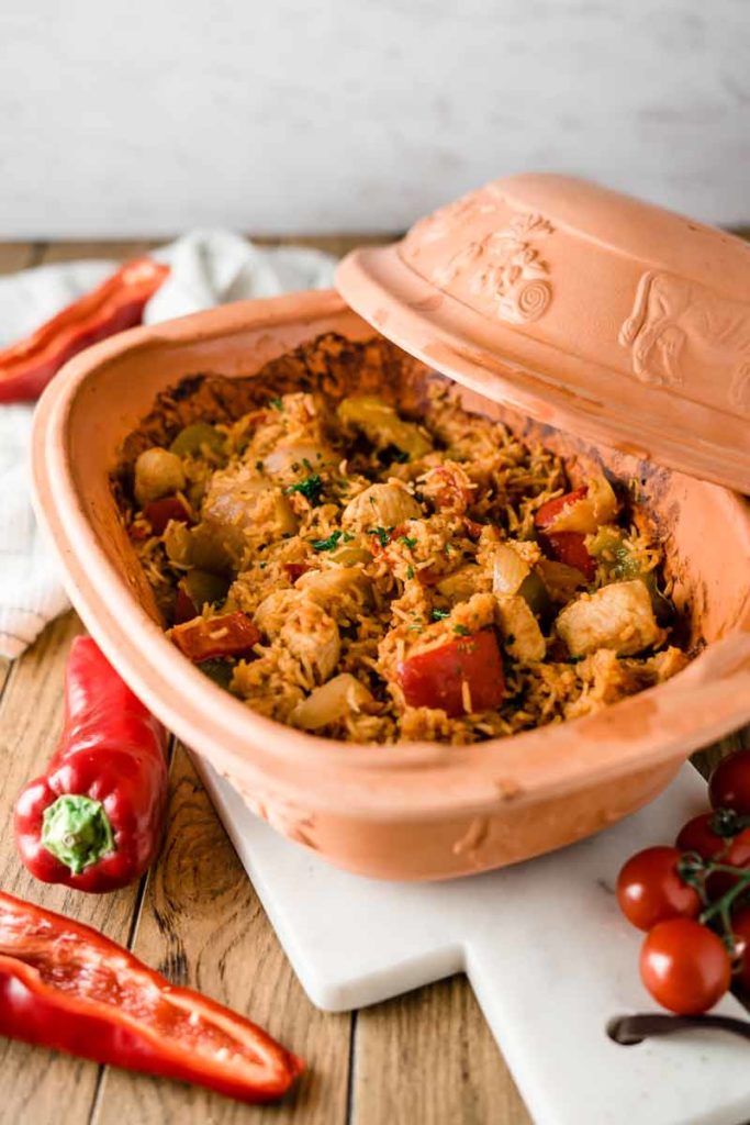 Keramiktopf mit Paprika-Hähnchen-Reis-Gericht, umgeben von frischen Paprikas und Tomaten auf Holzbrett.