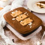 Tiramisu in einer Form, bestreut mit Kakao, verziert mit Keksen und Mandelsplittern, auf einem Tisch arrangiert.