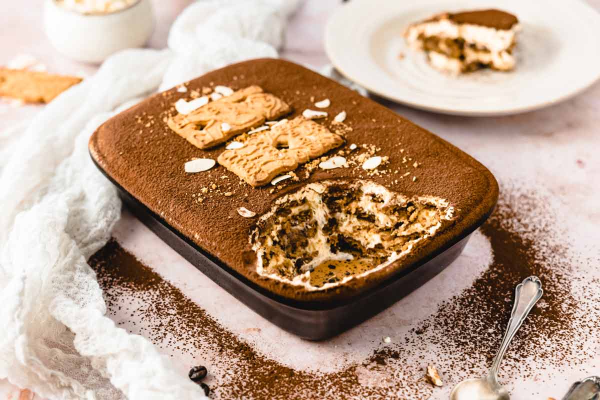 Tiramisu in dunkler Form, mit Löffel und zerbrochenen Keksen, auf rosa Hintergrund mit weißem Tuch.