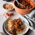 Tagliatelle mit Linsen-Bolognese in einer Schale, daneben ein Topf und Kirschtomaten auf einem Tisch.
