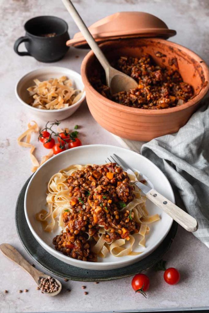Tagliatelle mit Linsen-Bolognese in einer Schale, daneben ein Topf und Kirschtomaten auf einem Tisch.