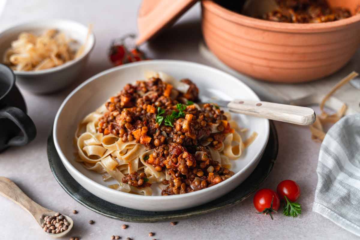 Tagliatelle mit Linsenbolognese in einem weißen Teller, umgeben von Zutaten auf einem Tisch.