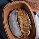 Ein gebackenes Brot in einem geöffneten Römertopf auf grauem Untergrund.