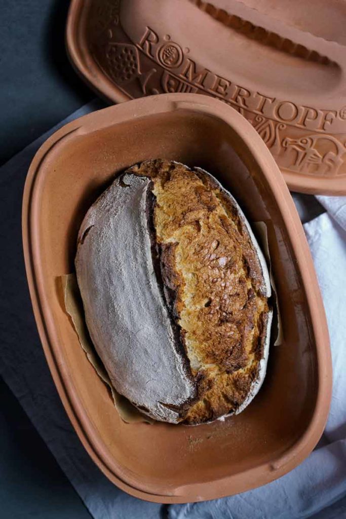Ein gebackenes Brot in einem geöffneten Römertopf auf grauem Untergrund.