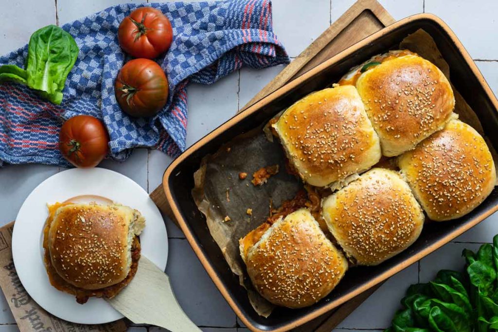 Frische Brötchen in einer Auflaufform, daneben Tomaten und Salatblätter auf einem blauen Tuch. Ein Brötchen auf Teller.