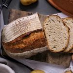 Brotlaib mit zwei Scheiben auf Holzbrett, umgeben von Kartoffeln und einem Römertopf auf dunkler Oberfläche.