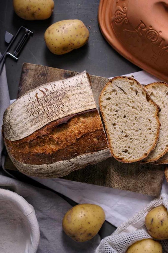 Brotlaib mit zwei Scheiben auf Holzbrett, umgeben von Kartoffeln und einem Römertopf auf dunkler Oberfläche.