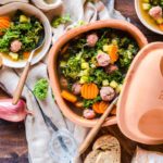 Eintopf mit Grünkohl, Kartoffeln, Möhren und Fleischbällchen in einem Tontopf, serviert mit Brot auf Holztisch.