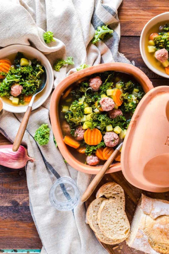 Eintopf mit Grünkohl, Kartoffeln, Möhren und Fleischbällchen in einem Tontopf, serviert mit Brot auf Holztisch.