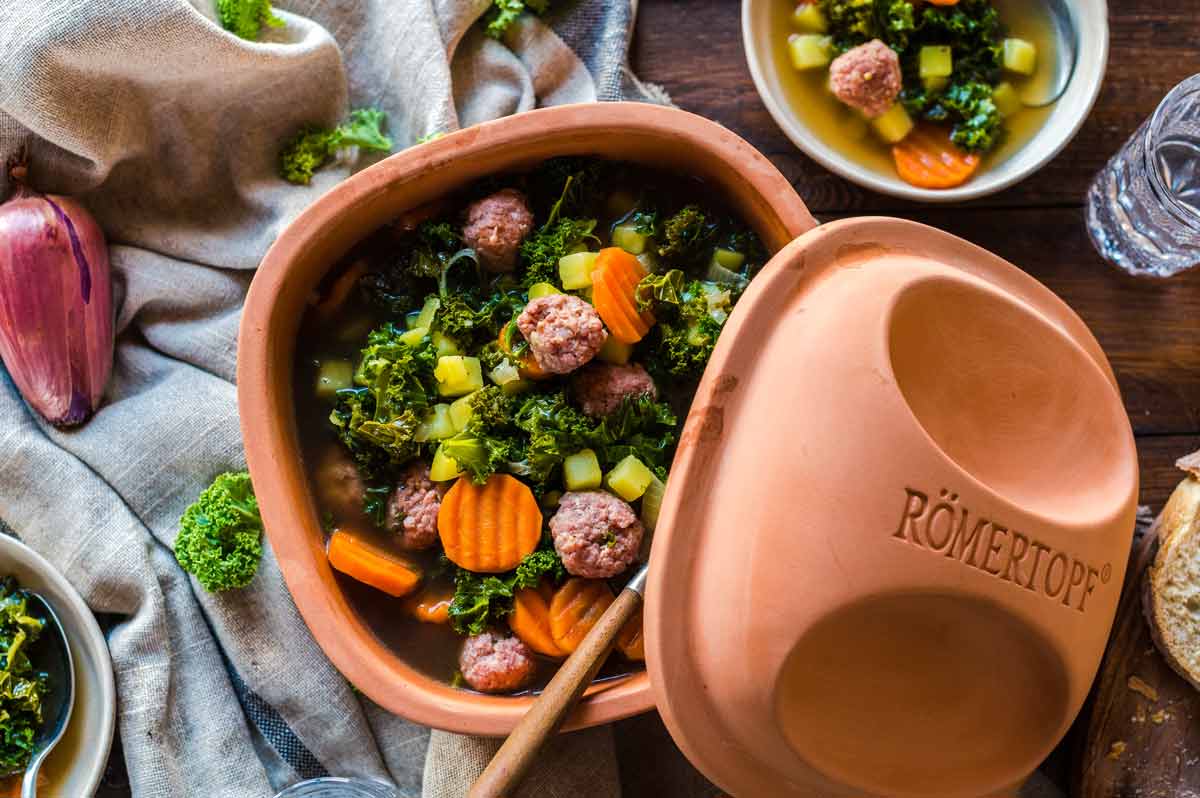 Römertopf mit Grünkohl, Möhren, Fleischbällchen und Kartoffeln, daneben eine Schüssel mit Suppe und ein Glas Wasser.
