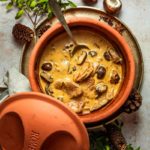 Ton-Topf mit Cremesuppe aus Fleisch und Pilzen, daneben Kräuter und Tannenzapfen.