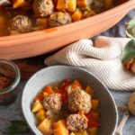 Eintopf mit Fleischbällchen, Kartoffeln und Karotten in einer Schüssel, im Hintergrund ein großer Terrakotta-Topf.