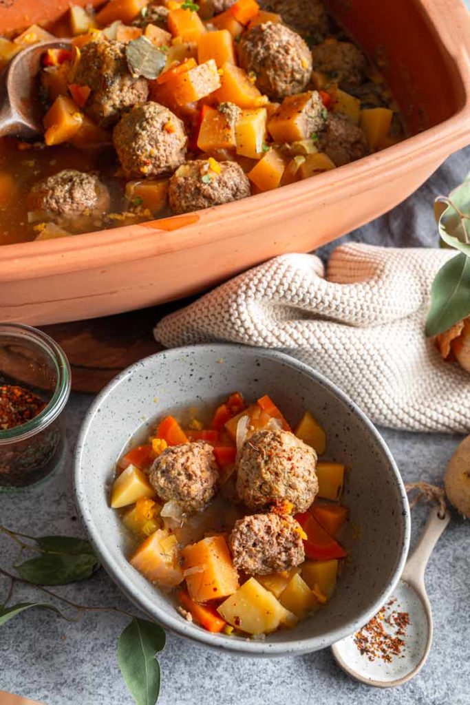 Eintopf mit Fleischbällchen, Kartoffeln und Karotten in einer Schüssel, im Hintergrund ein großer Terrakotta-Topf.