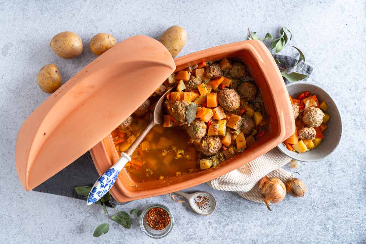 Auflauf im Tontopf mit Hackbällchen, Gemüse und Kartoffeln, dekoriert mit Gewürzen und Kräutern auf grauem Untergrund.