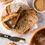 Rustikales Brot mit Butter aufgeschnitten, umgeben von Mehl, Getreide und einem Messer auf einem Tisch arrangiert.
