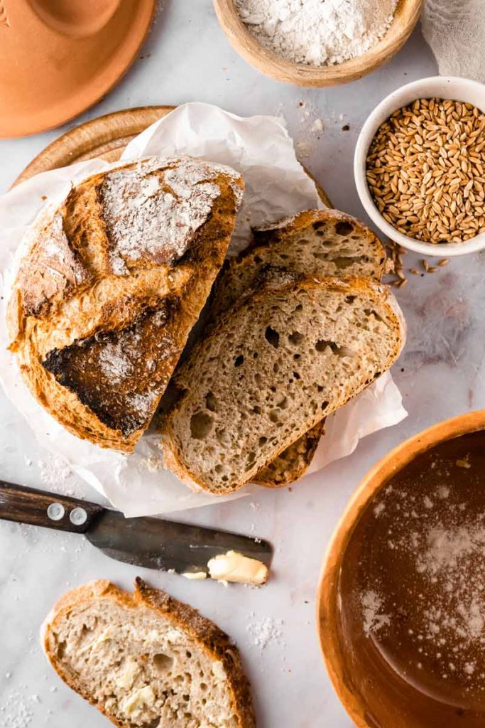 Rustikales Brot mit Butter aufgeschnitten, umgeben von Mehl, Getreide und einem Messer auf einem Tisch arrangiert.