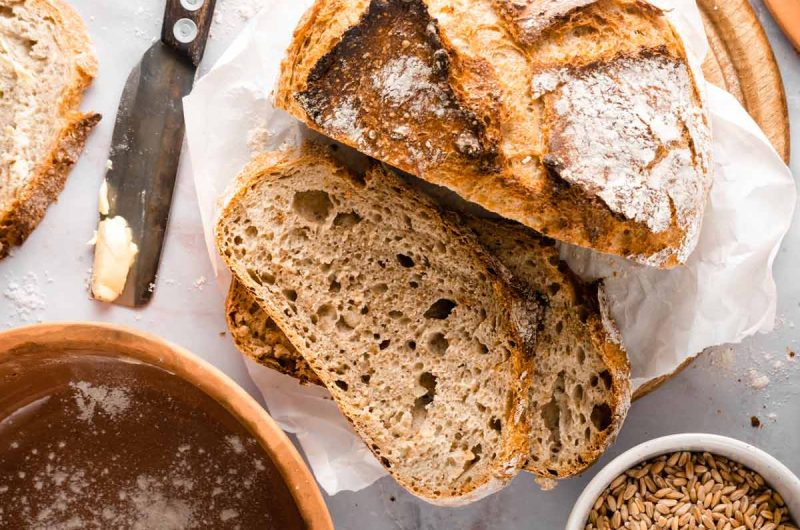 Frisch gebackenes Sauerteigbrot in Scheiben, daneben ein Messer mit Butter und eine Schüssel mit Getreidekörnern.