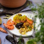 Teller mit Paella und Meeresfrüchten, garniert mit Kräutern. Im Hintergrund ein Topf mit mehr Paella.