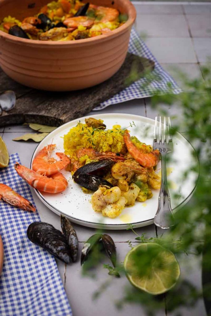 Teller mit Paella und Meeresfrüchten, garniert mit Kräutern. Im Hintergrund ein Topf mit mehr Paella.