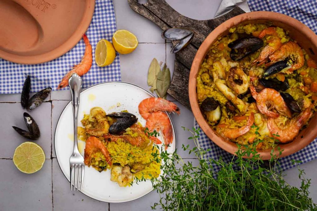 Paella mit Meeresfrüchten in einem Tontopf, daneben ein Teller mit Portion, Zitronen und Kräuter als Dekoration.