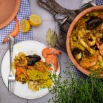 Paella mit Meeresfrüchten in einem Tontopf, daneben ein Teller mit Portion, Zitronen und Kräuter als Dekoration.