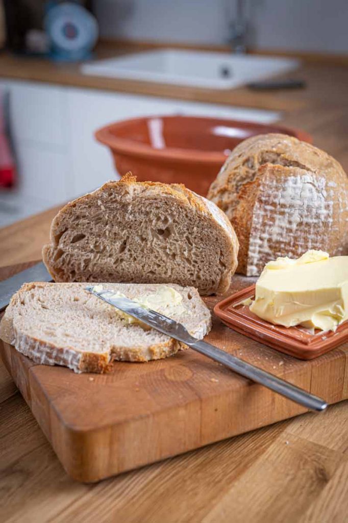 Scheiben hausgemachtes Brot mit Butter auf einem Holzbrett, daneben liegt ein Buttermesser.
