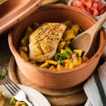 Hähnchenbrust auf Gemüsecurry in einem Tontopf, mit Holzlöffel und Gabel auf Holztisch angerichtet.