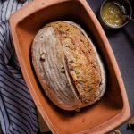 Hausgemachtes Brot in brauner Backform, umgeben von Walnüssen, Marmelade und gestreiftem Tuch auf dunklem Untergrund.