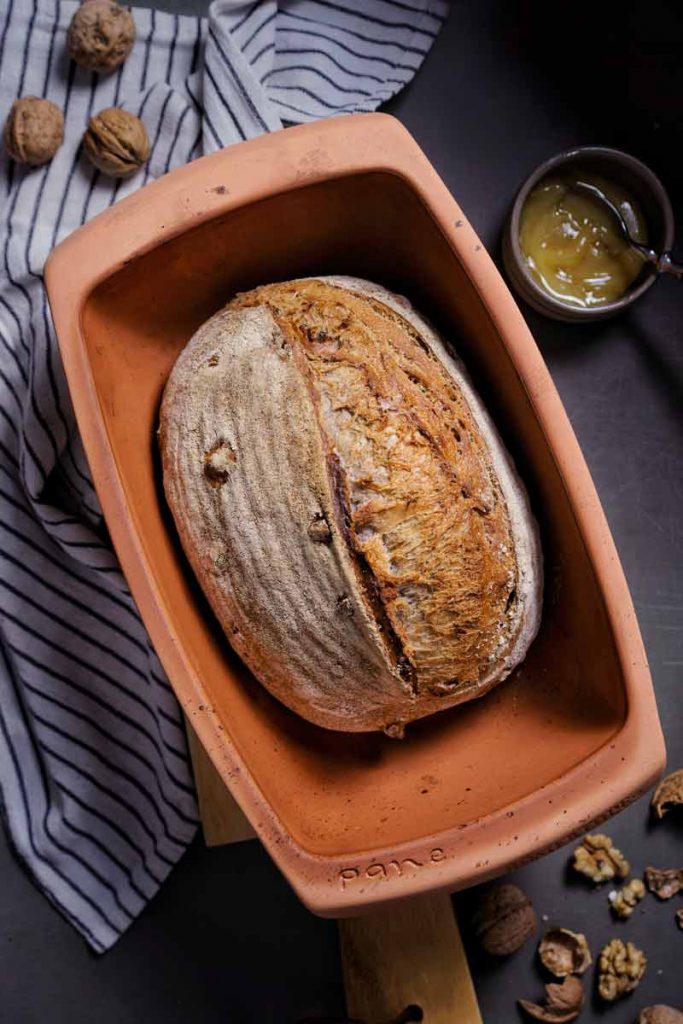 Hausgemachtes Brot in brauner Backform, umgeben von Walnüssen, Marmelade und gestreiftem Tuch auf dunklem Untergrund.